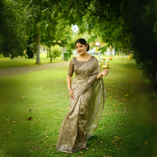 A woman models an elegant Beige Soft Cotton Saree featuring an all-over digital block print. She wears a heavily embellished, short-sleeve blouse in a matching beige/gold color and accessorizes with silver bangles, posing on a lawn under trees.