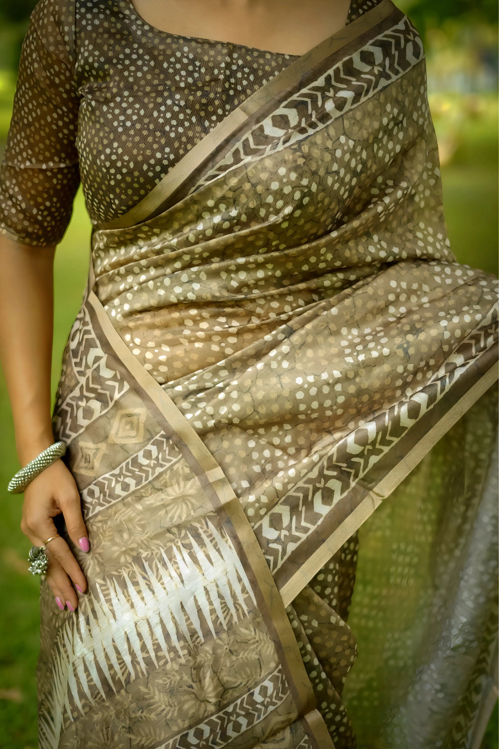 A woman models an elegant Beige Soft Cotton Saree featuring an all-over digital block print. She wears a heavily embellished, short-sleeve blouse in a matching beige/gold color and accessorizes with silver bangles, posing on a lawn under trees.