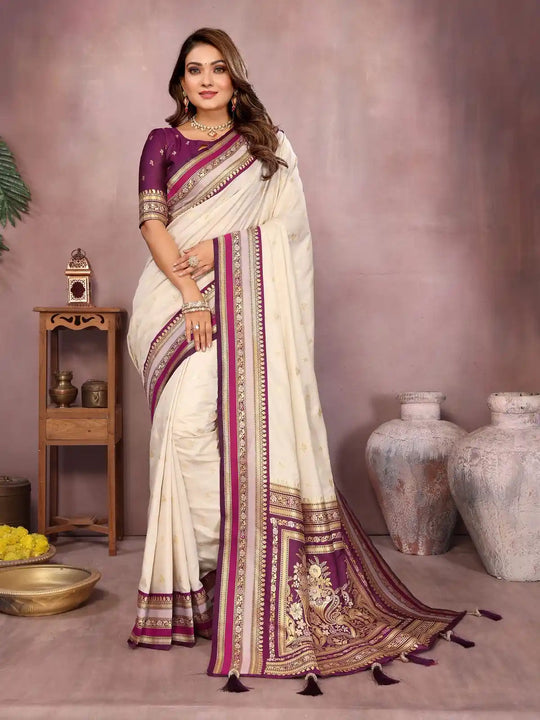 Woman wearing a white saree with purple and gold border in a decorated room.