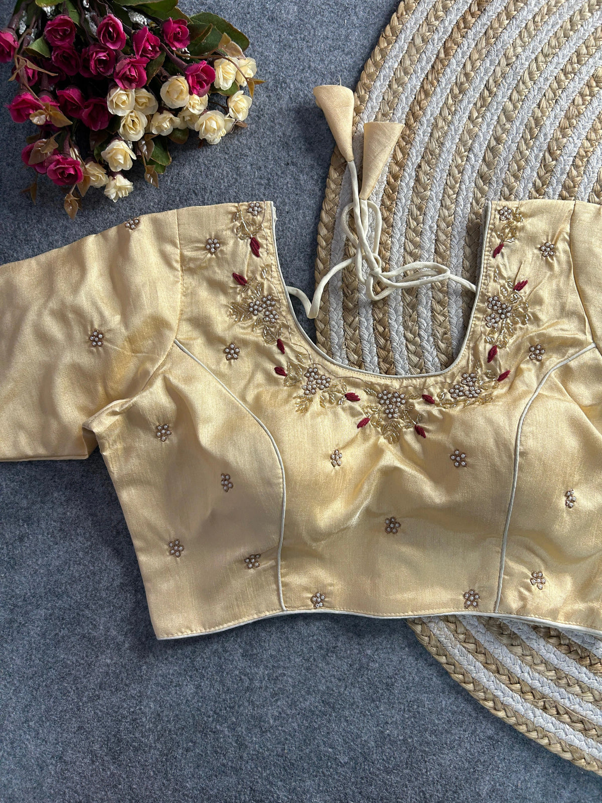 The front of the cream silk blouse features a curved V-neckline with matching pearl and gold floral handwork and maroon accents. The bodice is tailored with structured princess-line seams and adorned with scattered embroidered pearl motifs. The blouse is displayed on a grey textured surface next to a round jute mat and a bouquet of pink and white flowers.
