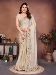 Woman in a cream saree with floral patterns standing in a decorated room.