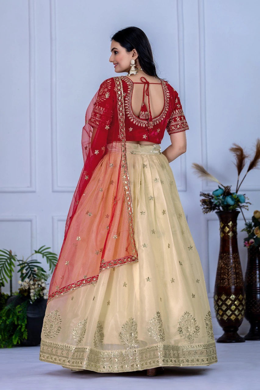 Woman wearing a red and beige traditional outfit with intricate designs against a white wall.