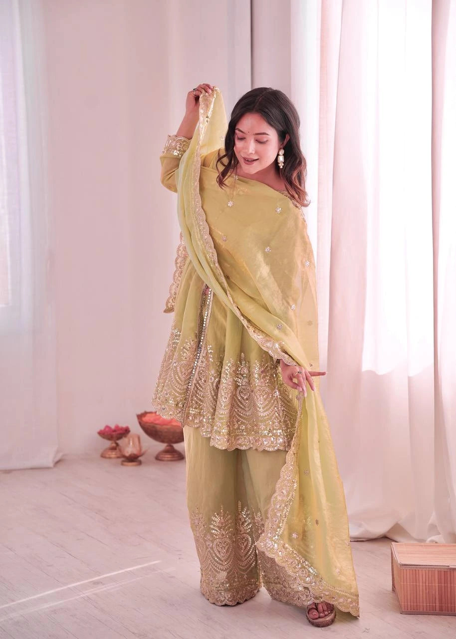 Woman in a light green traditional outfit with gold embroidery standing against a white curtain.