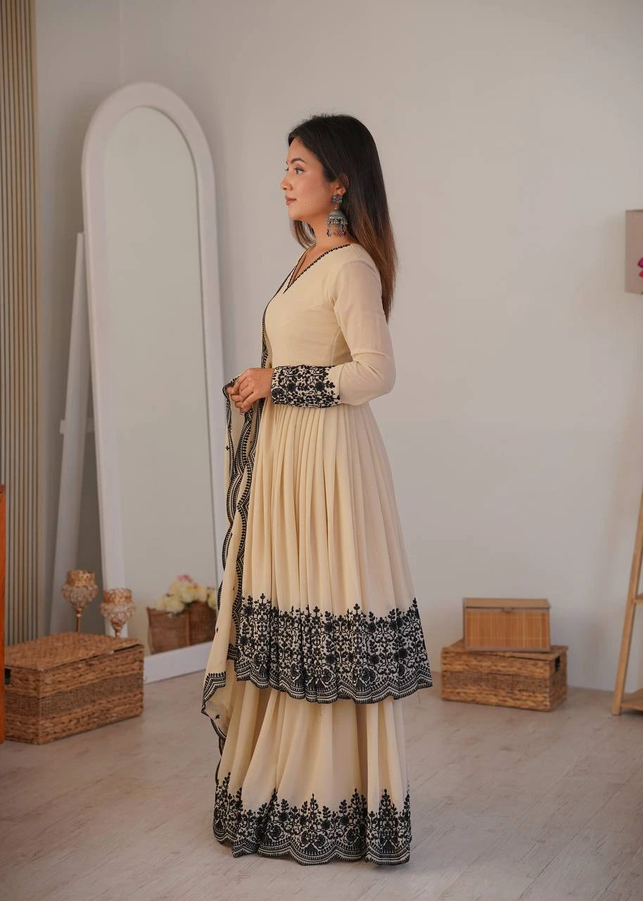 Woman in a cream and black traditional outfit standing in a room with wooden boxes and a mirror.