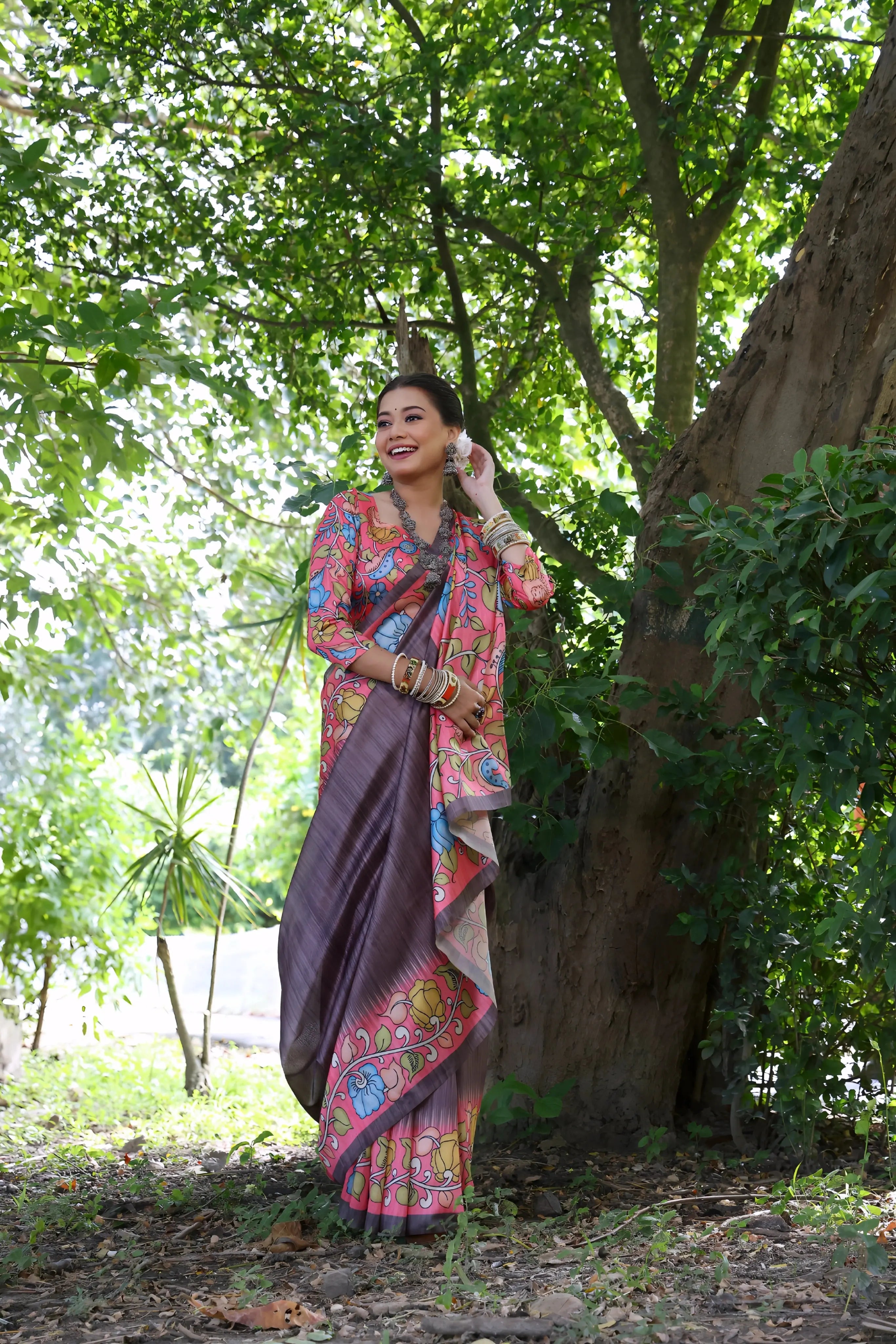 A woman models a unique Slate Grey/Smoky Plum Soft Tussar Silk Saree. The saree features a wide border and pallu with a vibrant Kalamkari-style print in Fuchsia Pink, sky blue, and green. She wears a matching long-sleeve printed blouse and accessorizes with heavy oxidized silver jewelry, including a layered necklace and stacked bangles, posing outdoors.