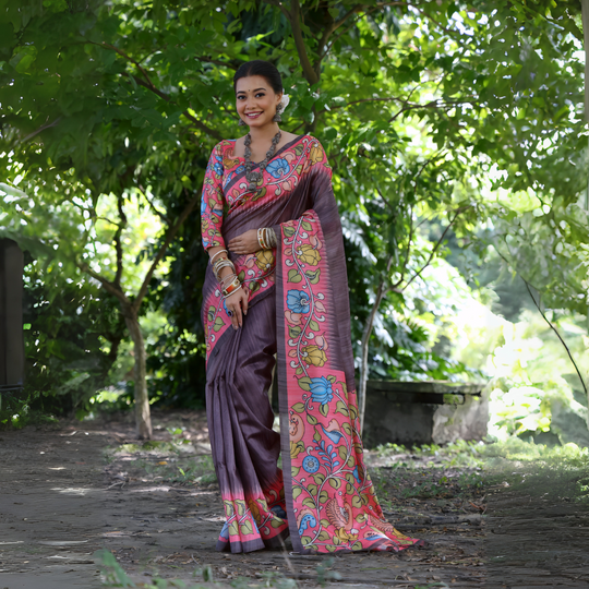 A woman models a unique Slate Grey/Smoky Plum Soft Tussar Silk Saree. The saree features a wide border and pallu with a vibrant Kalamkari-style print in Fuchsia Pink, sky blue, and green. She wears a matching long-sleeve printed blouse and accessorizes with heavy oxidized silver jewelry, including a layered necklace and stacked bangles, posing outdoors.