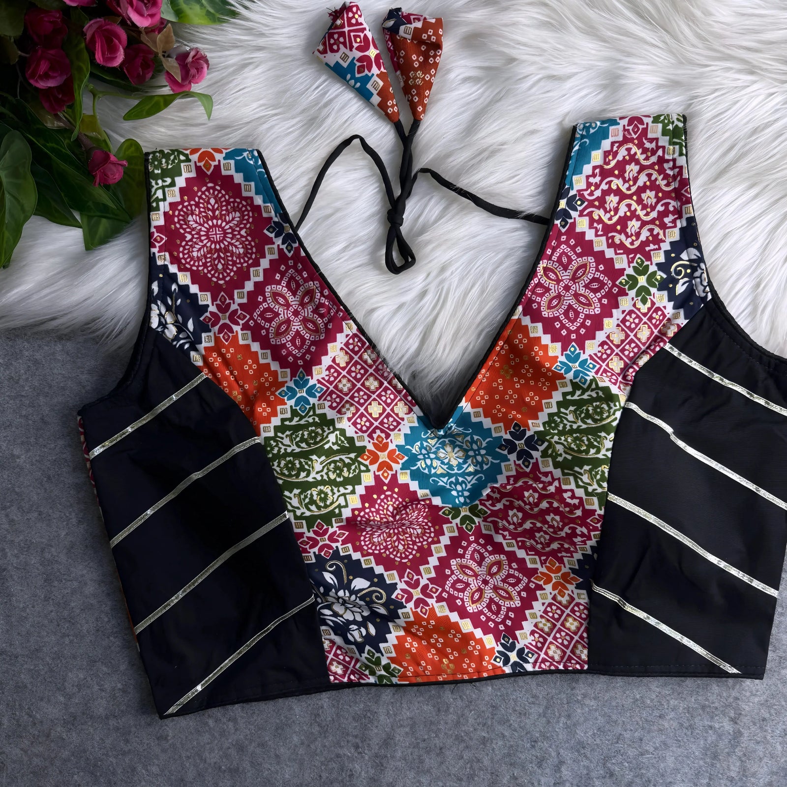 Traditional multicolor Ajrak-print blouse featuring vibrant geometric and floral motifs in orange, pink, blue, green, and red. The blouse has a deep V-neckline, black silk side panels with metallic silver stripes, and a patterned tasseled tie-back, displayed flat on a white fur background with artificial pink flowers on the side.