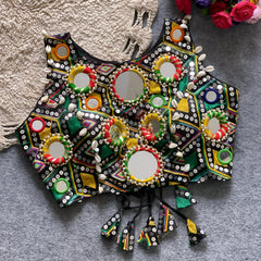 Traditional multicolor Kutch-style blouse featuring vibrant geometric mirror work surrounded by colorful beads accented with cowrie shells and tasseled tie-backs, displayed flat on a textured grey surface beside a braided cream mat and a bouquet of artificial roses.