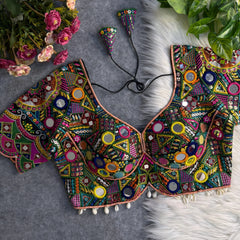 Traditional multicolor Kutch blouse featuring intricate mirror work, geometric embroidery, and vibrant threadwork in green, yellow, pink, blue, and orange. The design includes round and teardrop mirrors, beaded trim with cowrie shells, and tasseled tie-backs, displayed flat on a grey backdrop with artificial pink flowers and green foliage at the corners.