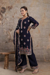 Woman wearing a navy blue embroidered traditional outfit against a textured wall.