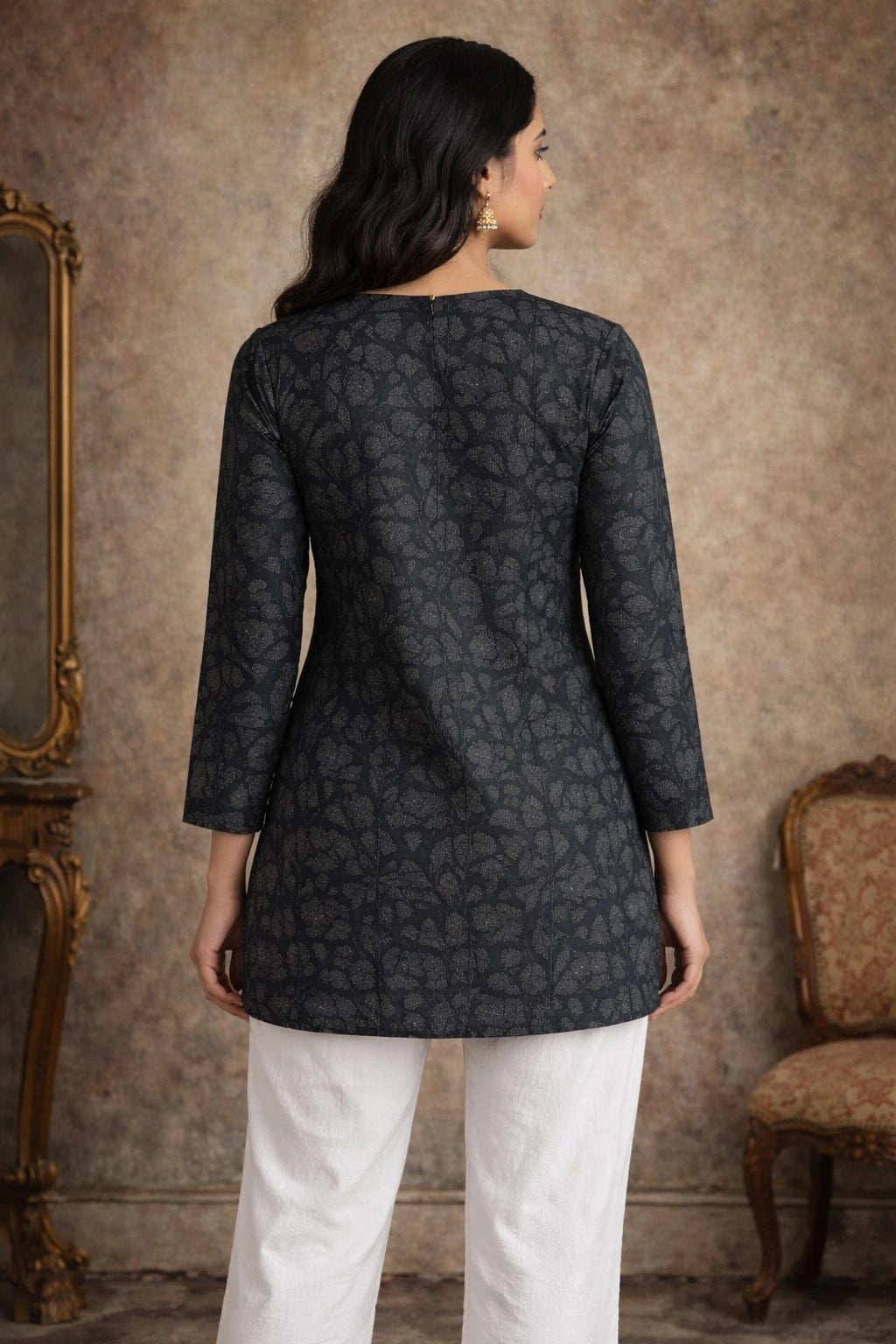 Woman wearing a dark patterned top and white pants against a textured wall.