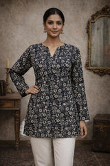 Woman wearing a dark floral patterned top with white pants in an indoor setting.