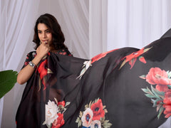 Woman wearing a black saree with floral patterns against a white curtain background