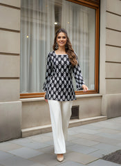 Woman in a black and white patterned top and white pants standing in front of a window.