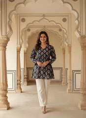 A woman stands facing forward in a black short kurti featuring a white and red ethnic block print. The top has a V-neckline, long sleeves, and a gathered empire waist that flares into a peplum hem. She is wearing off-white trousers and large silver jhumka earrings.