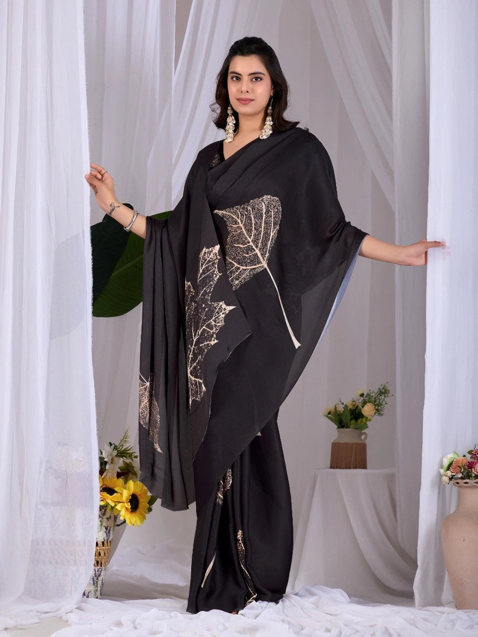 Woman wearing a black saree with floral patterns against a white curtain background