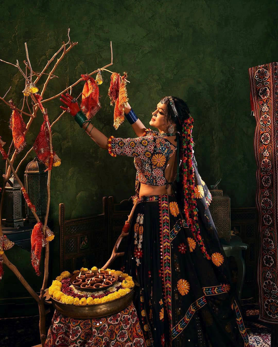 Woman in traditional attire performing a ritual with offerings in a decorated room.