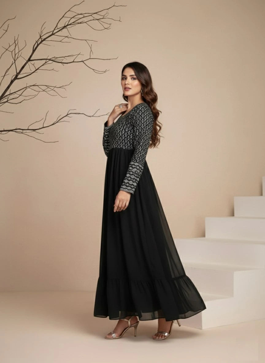 Woman in a black and patterned long dress standing on a staircase with a beige wall and branch in the background.
