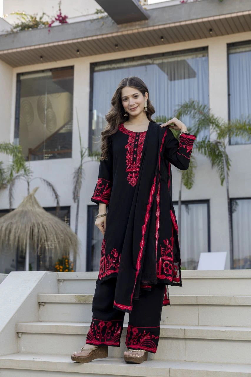 Woman wearing a black and red embroidered outfit standing on steps with a building and plants in the background.