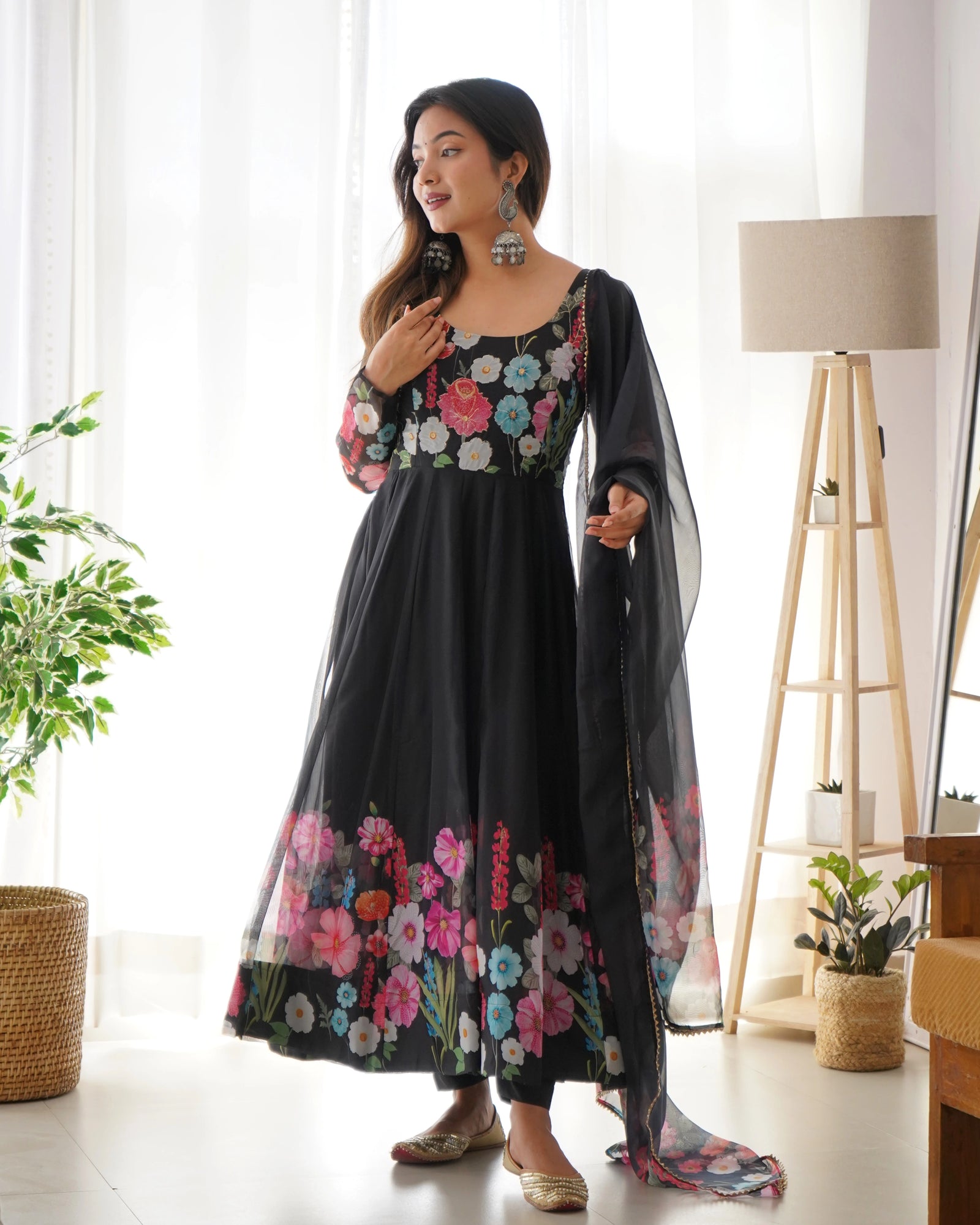 Woman wearing a black floral outfit in a bright room with plants and a lamp.
