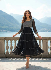 Woman in a black dress with a patterned top standing by a scenic lakeside.