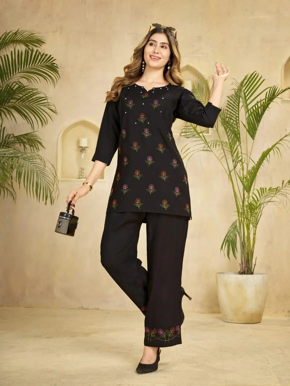 Woman wearing a black traditional outfit with floral patterns in an indoor setting with plants.