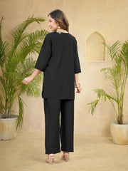 Woman in a black outfit standing between two potted plants in a room with beige walls.