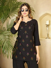 Woman wearing a black embroidered top in an indoor setting with a plant and candle in the background.