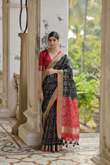 Woman in a black and red saree standing in an outdoor setting with greenery.