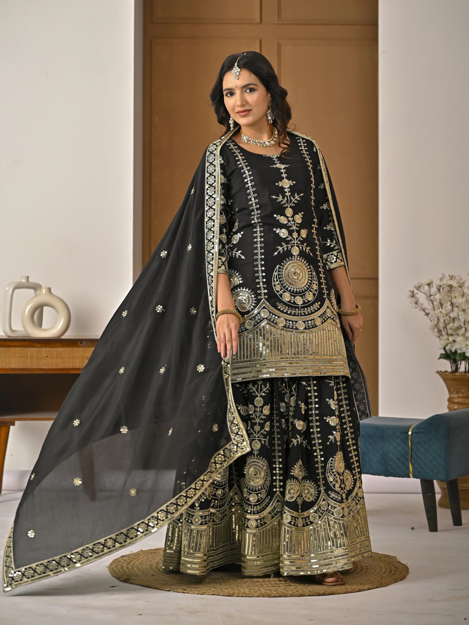 Woman in traditional black and gold embroidered outfit with a sheer dupatta in a room.