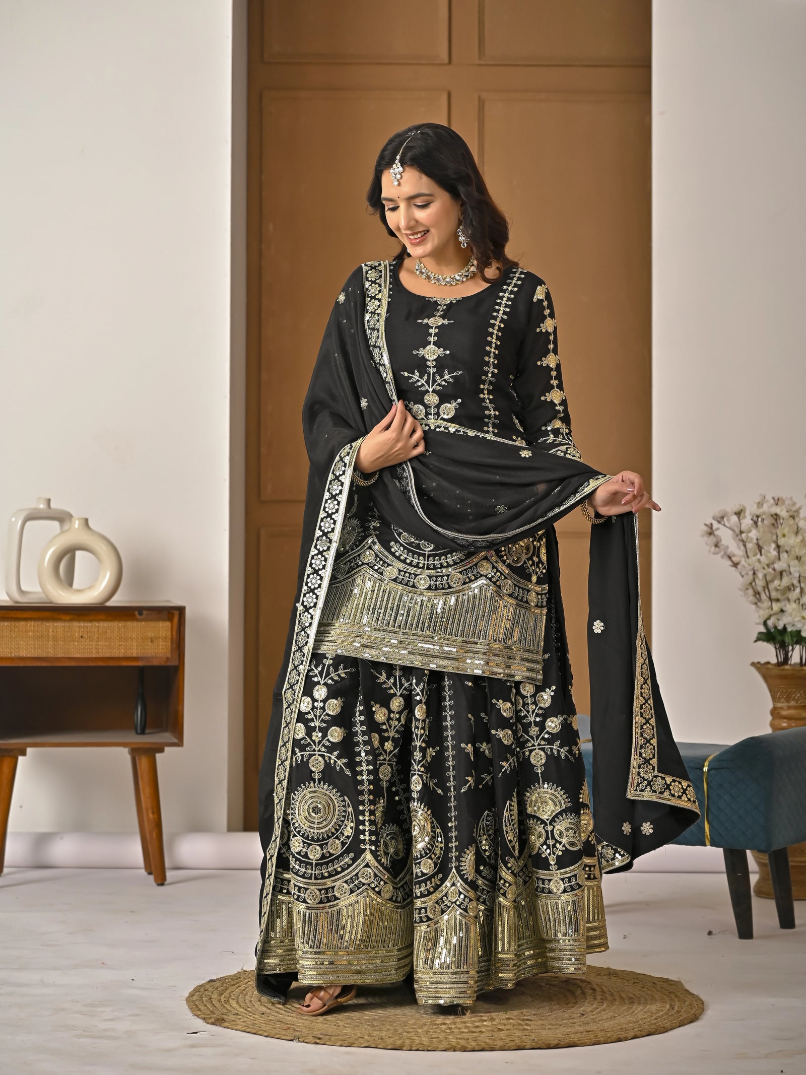Woman in traditional black and gold embroidered outfit standing in a room with wooden furniture.