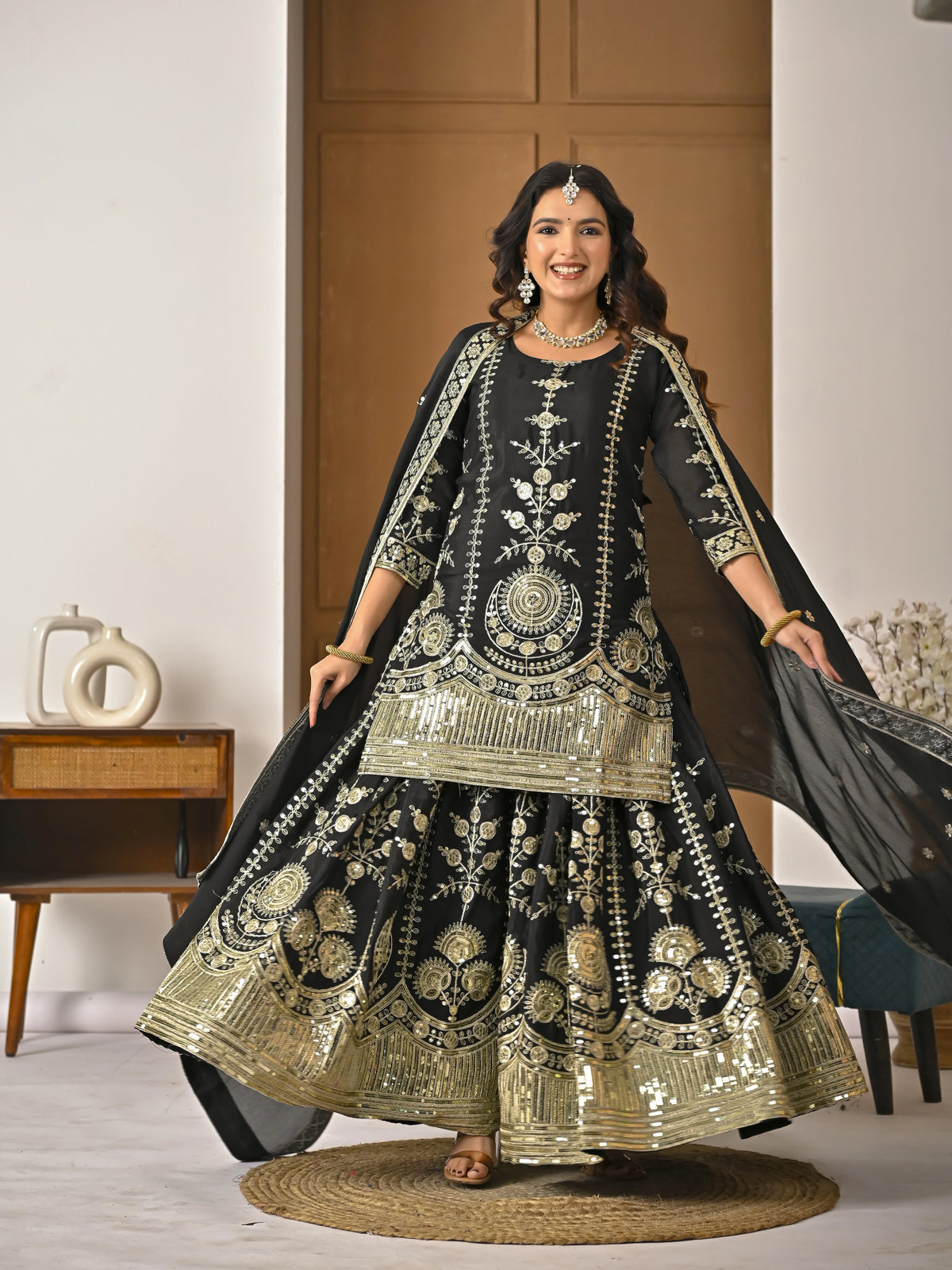 Woman in a black and gold embroidered traditional outfit standing indoors.