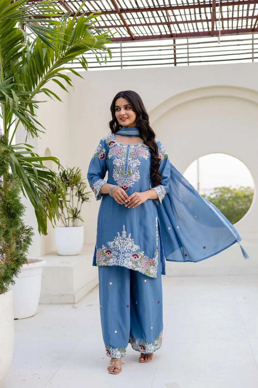 Woman in a blue embroidered traditional outfit standing in a bright outdoor setting with plants.