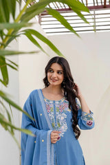 Woman in a blue embroidered outfit standing outdoors with greenery.