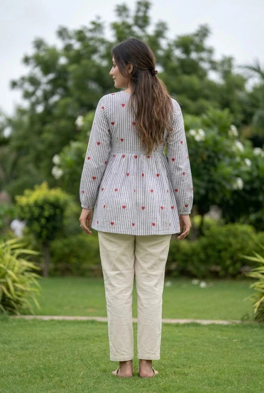 Woman standing outdoors wearing a patterned top and beige pants with greenery in the background