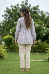 Woman standing outdoors wearing a patterned top and beige pants with greenery in the background