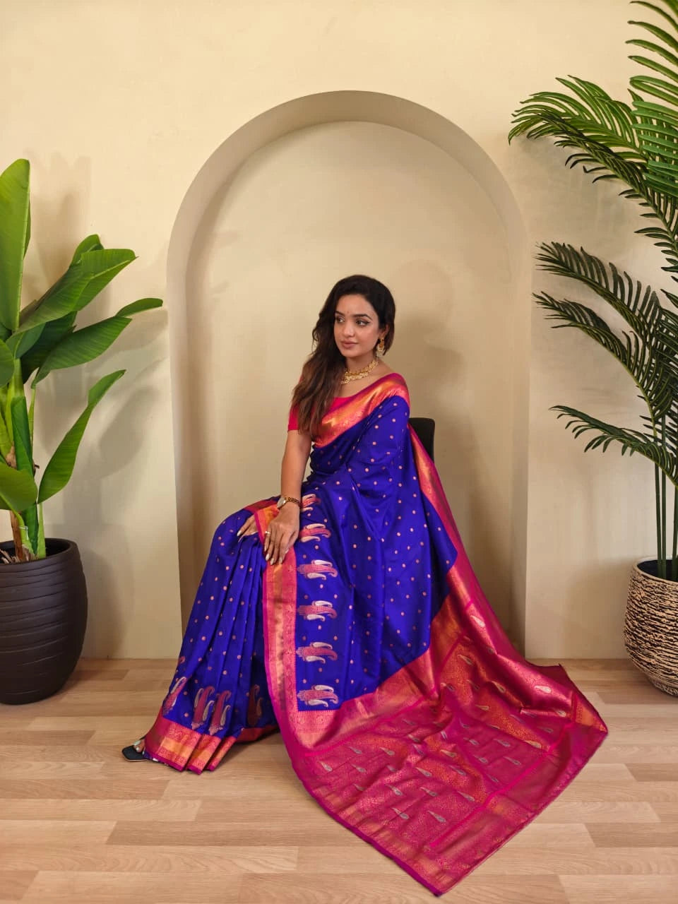Woman in a blue and pink saree sitting indoors with plants around