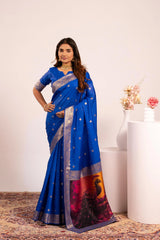 Woman wearing a blue saree with a scenic design, standing against a white wall.