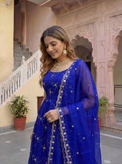 Woman in a blue traditional outfit with gold embroidery standing in front of an architectural building.