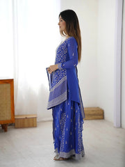 Woman in a blue traditional outfit holding a matching bag in a minimalistic room.