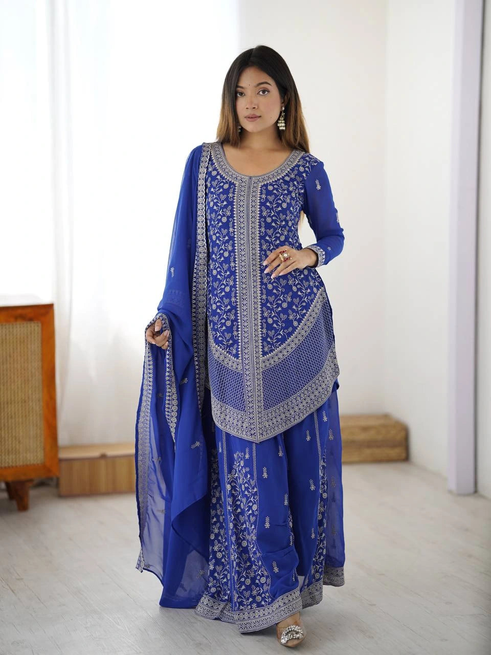Woman wearing a blue embroidered traditional outfit in a minimalistic room.