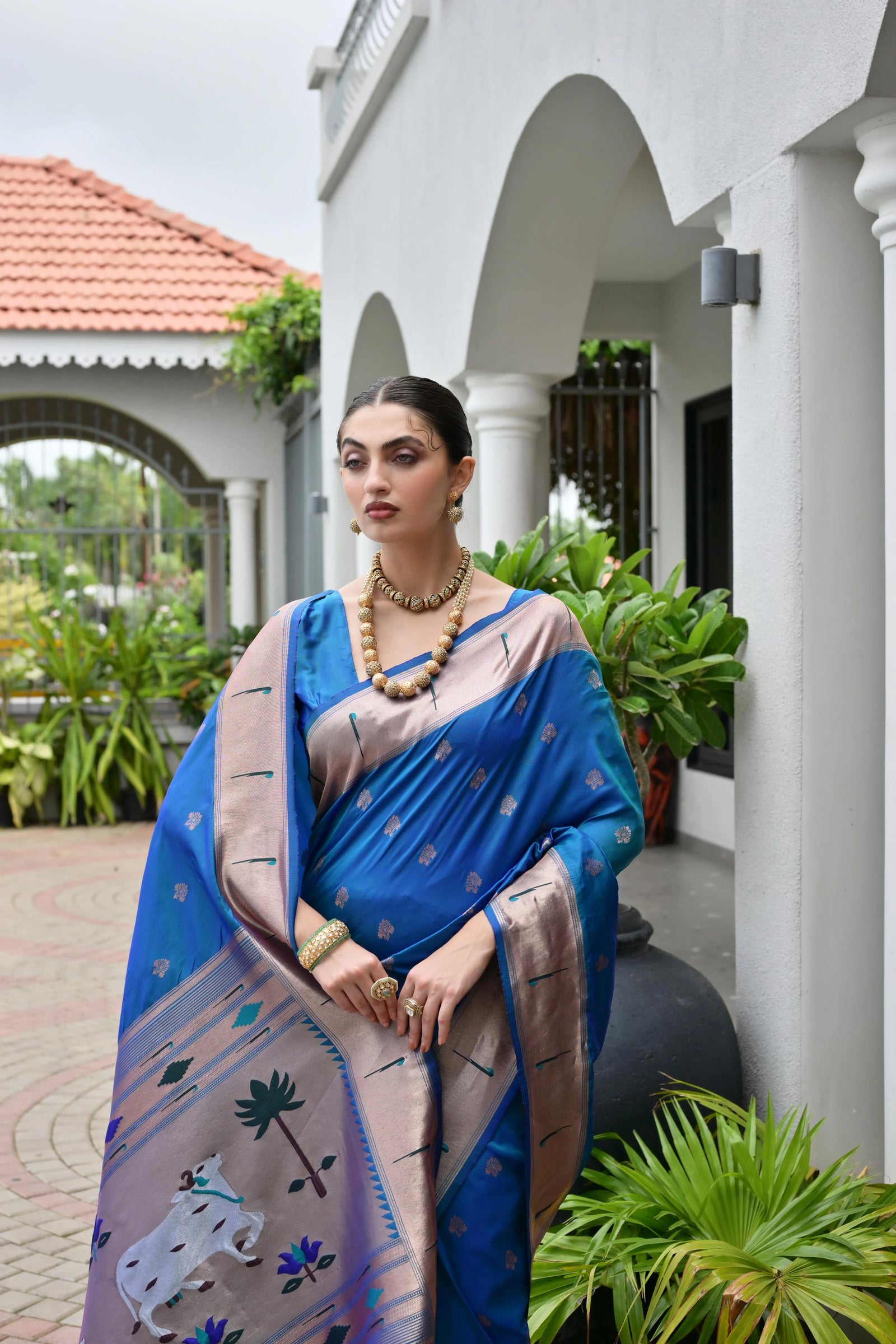 Woman in a blue saree with a white building and plants in the background