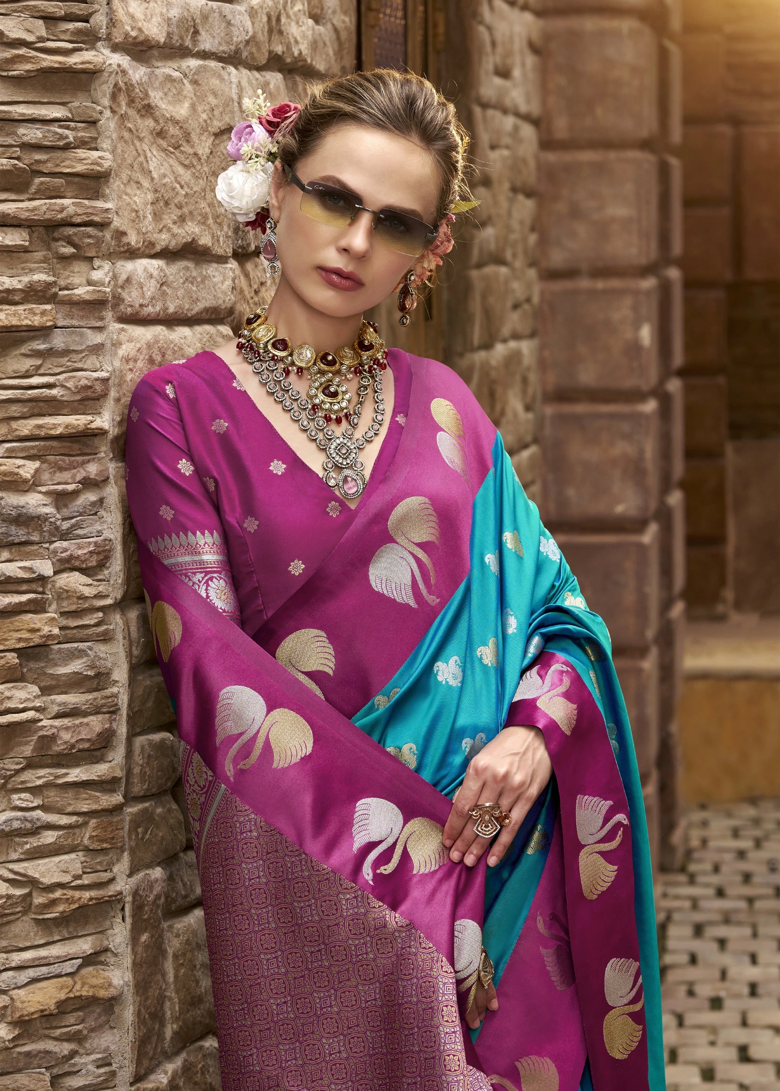 Woman in a traditional pink and blue saree leaning against a stone wall.
