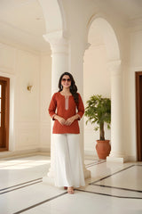 Woman in a red top and white pants standing in a sunlit hallway with columns and a plant.
