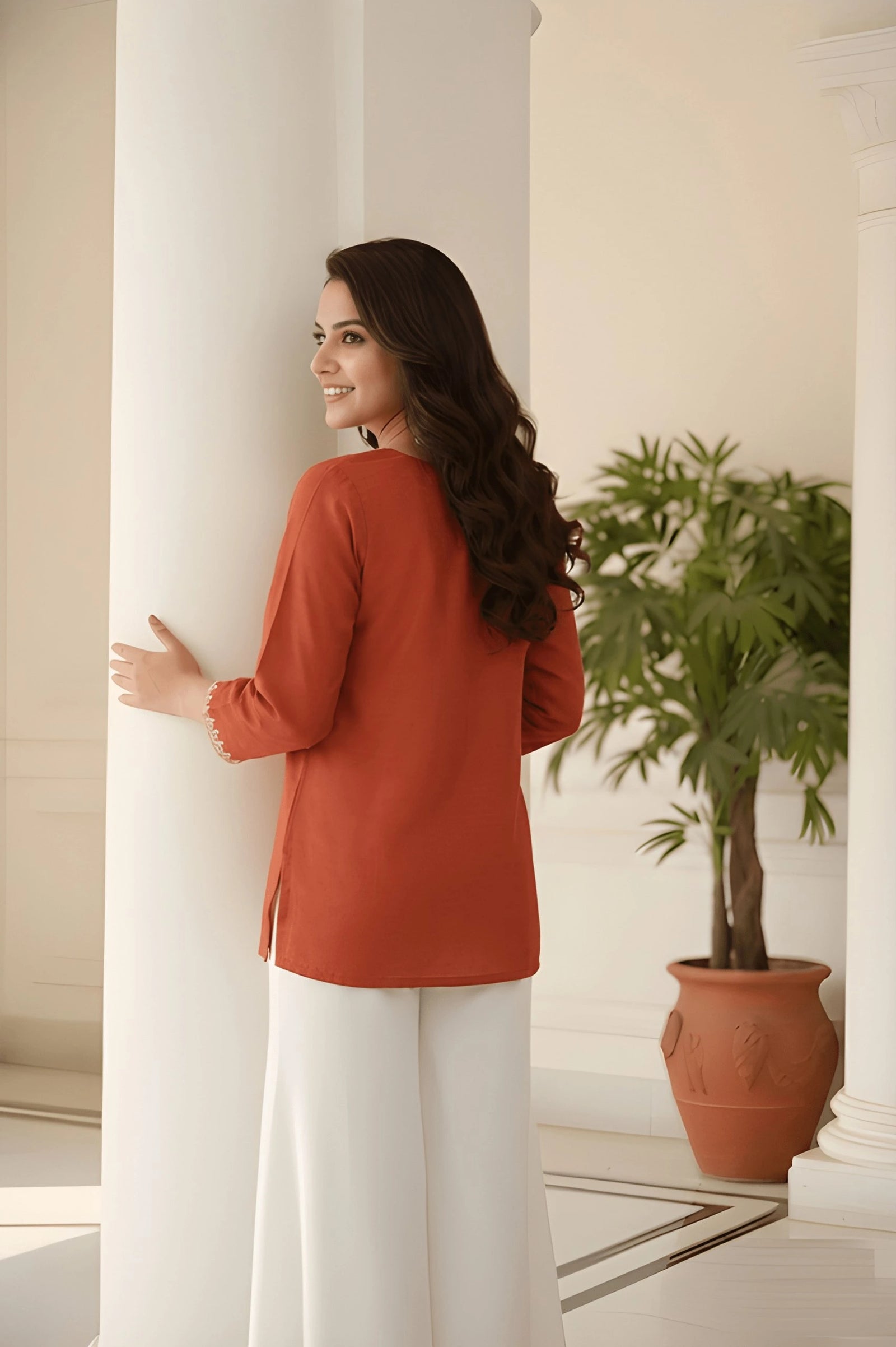 Woman in a red top and white pants standing in a room with a plant and columns.