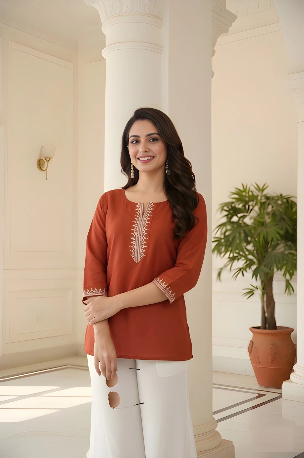 Woman wearing a rust-colored top with white pants standing in a well-lit room with columns and a plant.