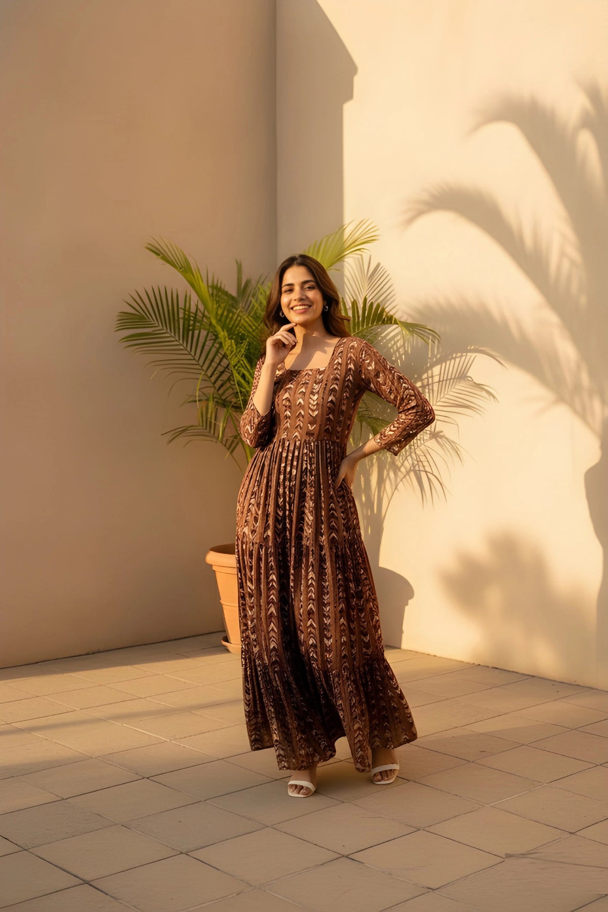 Woman in a long, patterned dress standing against a light wall with a plant.