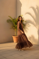 Woman in a long, patterned dress standing in front of a light-colored wall with a potted plant.