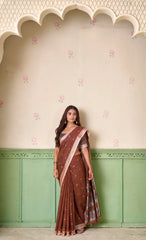 Woman wearing a brown saree with a pink border against a decorative wall.