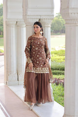 Woman in traditional brown outfit standing in front of a white architectural structure with greenery in the background